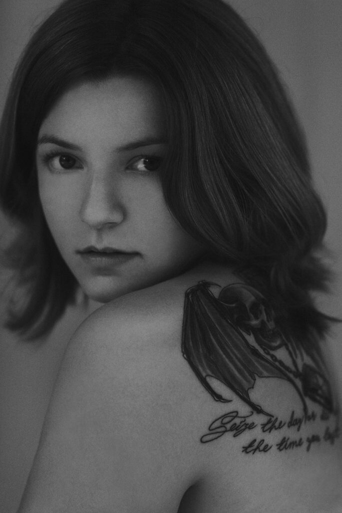 Black and white Portrait of a girl with tattoo on her shoulder; death's head with wings; Seize the day or die regretting the time you lost