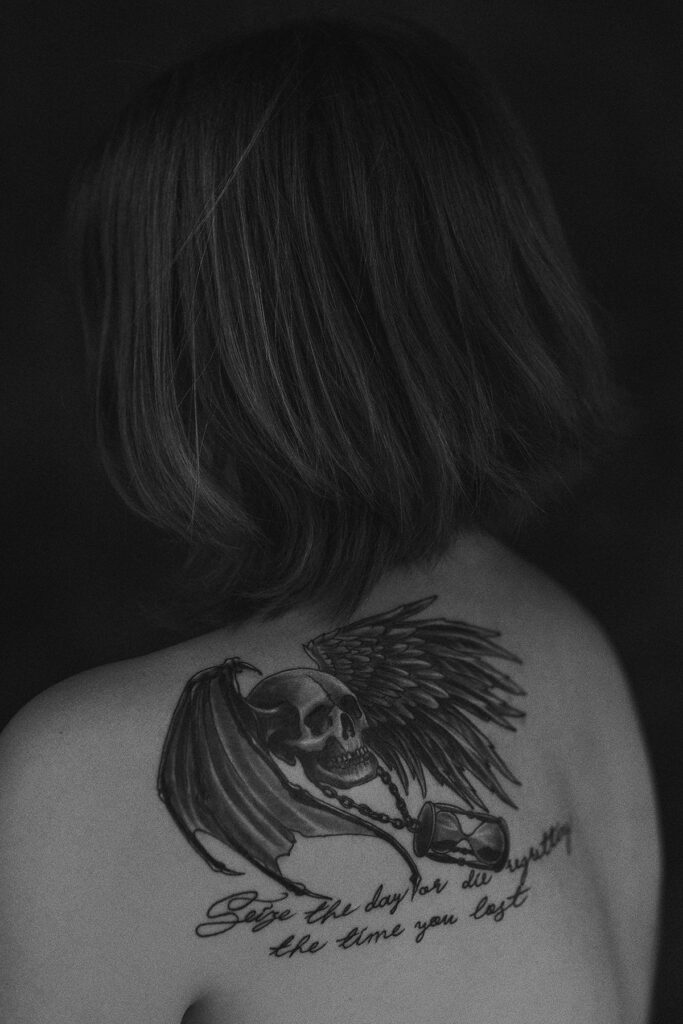 Black and white Portrait of a girl with tattoo on her shoulder; death's head with wings; Seize the day or die regretting the time you lost